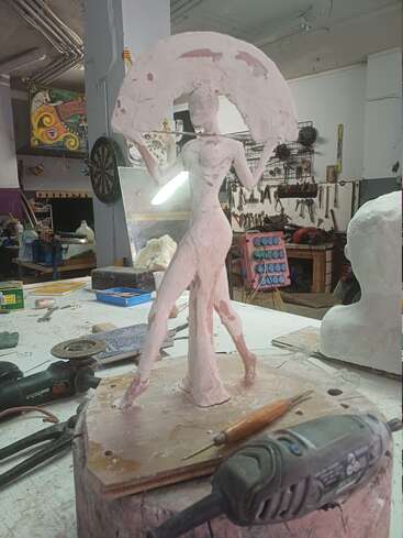 Una escultura de arcilla de una mujer que sostiene un paraguas sobre un banco de trabajo, rodeada de herramientas de tallado y equipos de taller en el estudio de un artista.