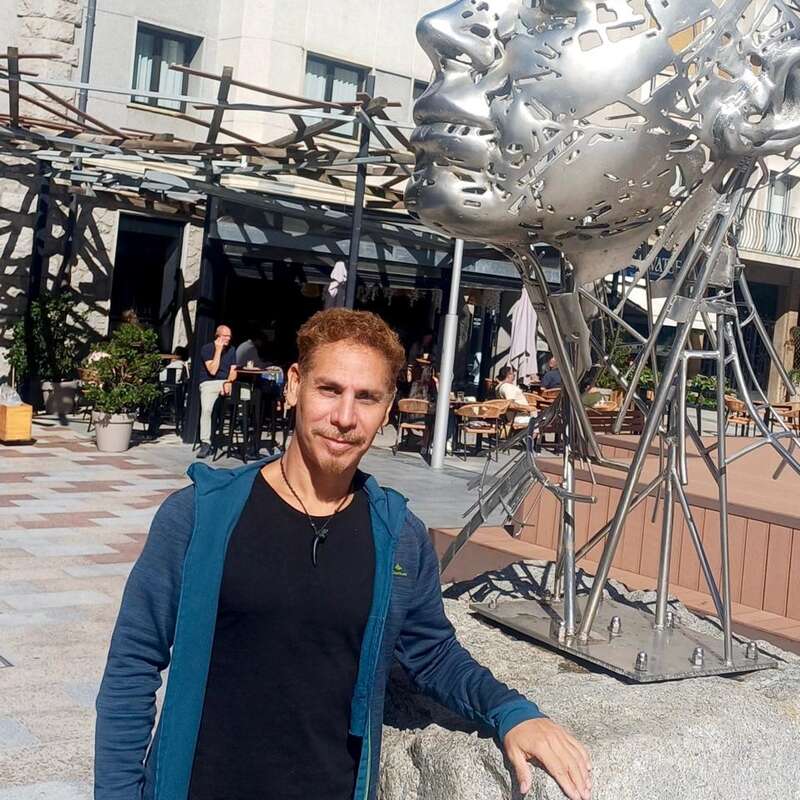 Un hombre junto a una gran escultura abstracta metálica de una cabeza al aire libre. El entorno soleado incluye un edificio moderno y una cafetería con gente sentada.