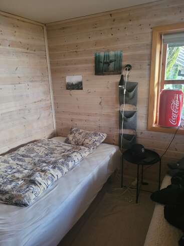 Una acogedora habitación con paneles de madera, una cama individual, ropa de cama estampada, una pequeña mesa redonda, cuadros en las paredes, un organizador colgante, una ventana y una nevera roja de Coca-Cola.