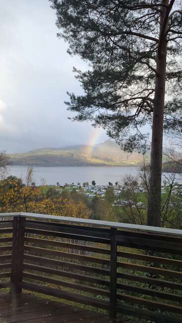 Una vista panorámica desde un balcón con barandillas de madera da a un lago, árboles otoñales, cabañas lejanas, montañas y un hermoso arco iris que brilla a través de un cielo nublado.
