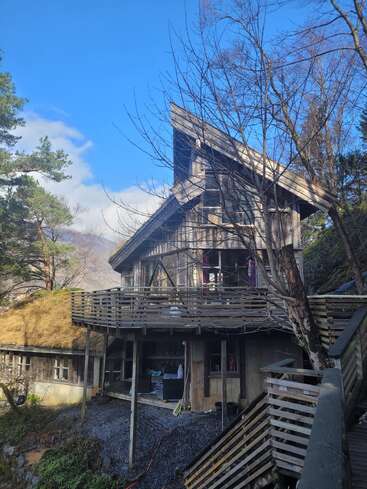 Esta imagen muestra una casa rústica de madera con un techo angular empinado, rodeada de árboles, cielo azul y montañas, con una gran terraza y una escalera exterior.