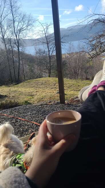 Una persona se relaja al aire libre con un café en la mano, las piernas estiradas y un perro a su lado, disfrutando de una vista panorámica de árboles, colinas y un lago.