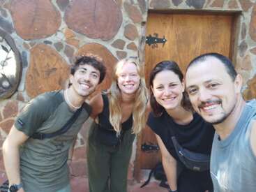 Four smiling friends pose together outside a rustic building with a wooden door and stone walls, enjoying a bright day and appearing happy and relaxed in each other's company.