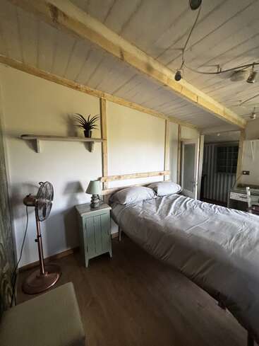 This cozy bedroom features a neatly made bed, wooden nightstand with a lamp, floor fan, potted plant on a shelf, and rustic decor with wooden accents.