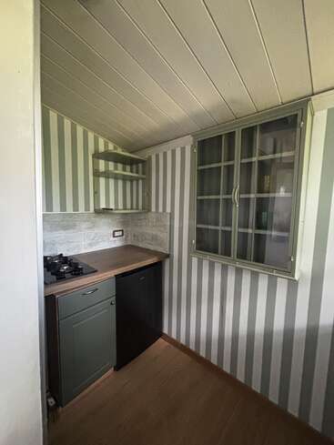 This small kitchen features striped wallpaper, a slanted ceiling, compact gas stovetop, countertop, under-counter fridge, open shelves, and glass-fronted wall cabinet with wooden flooring.