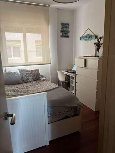 A cozy small bedroom with a bed, desk, and chest of drawers. Decor includes motivational signs, plants, and a laptop, with natural light from the window.