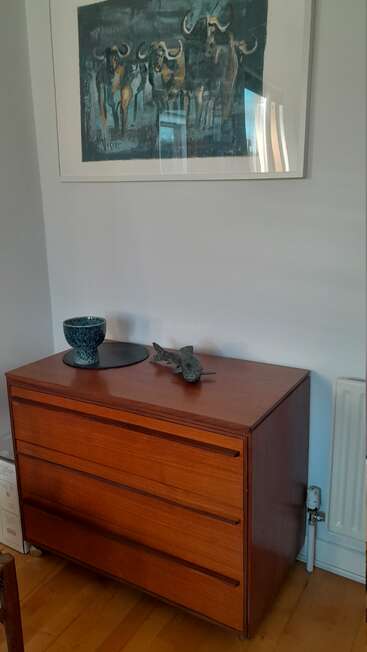 Une commode en bois est adossée au mur, décorée d'un bol en céramique, d'une sculpture de poisson et d'un tableau représentant des gnous au-dessus.