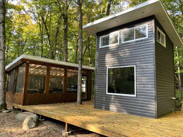 Ein modernes kleines Haus im Wald mit großen Fenstern und einem schrägen Dach, das an eine abgeschirmte Veranda grenzt und auf einem geräumigen Holzdeck steht.
