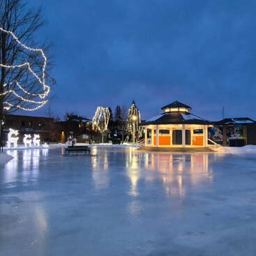 Eine bezaubernde Winterszene mit einer gefrorenen Schlittschuhbahn, einem beleuchteten Pavillon, geschmückten Bäumen, leuchtenden Rentierfiguren, Bänken und einem friedlichen, dunkelblauen Himmel.