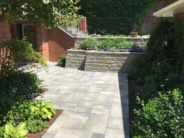 Dieses Bild zeigt einen wunderschön gestalteten Garten mit Steinfliesen, erhöhten Pflanzkübeln aus Ziegelsteinen, die mit Blumen bepflanzt sind, üppigem Grün und einer Treppe, die zu einer höheren Ebene führt.