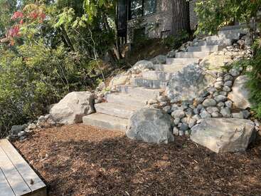 Steinstufen führen einen abfallenden Garten hinauf, der von großen Felsen und kleineren Kieselsteinen gesäumt ist. Bäume und Grünflächen umgeben den Bereich und führen zu einem Backsteinhaus.