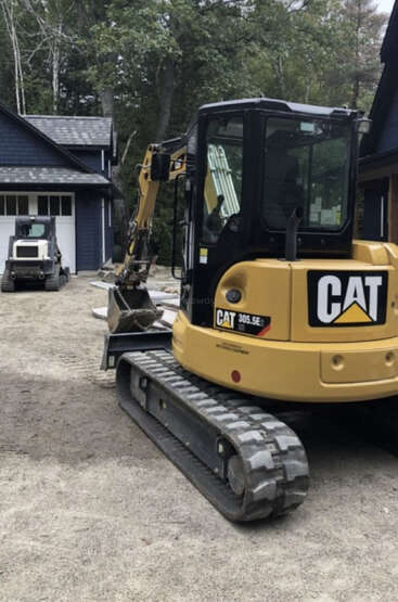 Ein gelber CAT 305.5E Minibagger steht auf unbefestigtem Boden in der Nähe eines Hauses. Im Hintergrund steht ein weiteres kleines Baufahrzeug neben einer Garage. Die Umgebung ist von Bäumen umgeben.