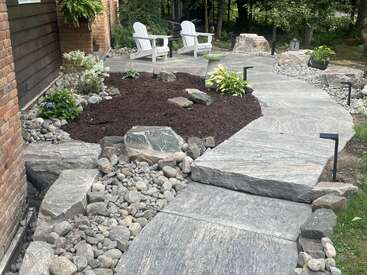 Ein Steinweg führt durch einen Landschaftsgarten mit Mulch, Steinen und Pflanzen. Auf einer Terrasse stehen zwei weiße Adirondack-Stühle, die zum Entspannen im Grünen einladen.