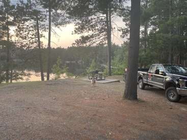 Ein ruhiger Campingplatz am See mit einem Picknicktisch, einem Hund, der daneben steht, einem geparkten schwarzen Pick-up, umgeben von hohen Kiefern bei Sonnenuntergang.