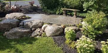 Eine friedliche Gartenszene mit großen Felsen, grünen Sträuchern und blühenden Pflanzen. Auf einer Terrasse steht eine Steinbank, umgeben von üppigem Grün und Sonnenlicht.