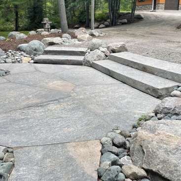 Auf diesem Bild sind große Steinstufen und ein Steinweg zu sehen, die von Felsen und Bäumen umgeben sind und zu einer Schotterauffahrt und einem Gebäude im Hintergrund führen.