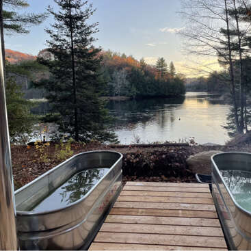 Dos bañeras metálicas llenas de agua se asientan sobre una terraza de madera con vistas a un lago sereno rodeado de árboles, bajo un cielo despejado al atardecer. Ambiente apacible.
