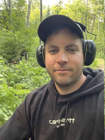 A man wearing a black Carhartt hoodie, black cap, and ear protection is outdoors in a green forest, taking a selfie with a slight smile.