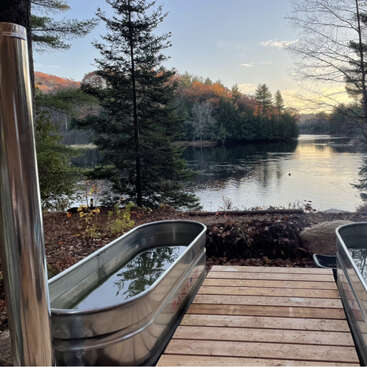 Two metal tubs filled with water sit on a wooden deck overlooking a serene lake surrounded by trees, under a clear sky at sunset. Peaceful atmosphere.