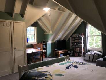 This cozy attic room features sloped ceilings, a bed, bookshelves, TV, reading nook, desk by the window, and soft natural light through large windows.