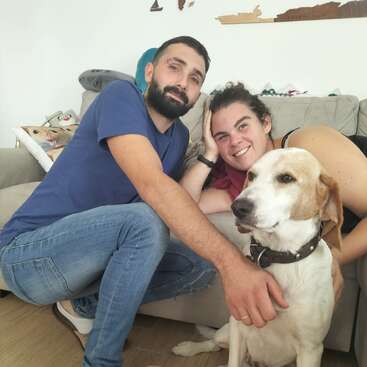 Das Bild zeigt einen Mann und eine Frau, die mit einem Hund auf einer Couch sitzen und für ein Foto posieren, umgeben von ein paar Kissen und einer weißen Wand im Hintergrund.