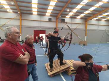 Dans un gymnase, plusieurs personnes participent à une épreuve de tir à l'arc. Certains tirent des flèches, tandis que d'autres observent attentivement. L'atmosphère semble concentrée et compétitive.
