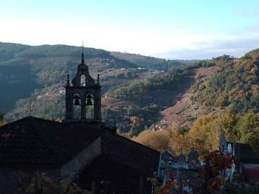 Un village pittoresque situé au sommet d'une colline présente un clocher d'église rustique au premier plan, entouré d'arbres luxuriants et colorés et de montagnes ondulantes et verdoyantes baignées de lumière.