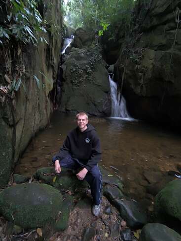 Un homme vêtu de sombre est assis sur des rochers moussus près d'une petite cascade dans une forêt luxuriante et verdoyante. La scène est sereine et naturelle.