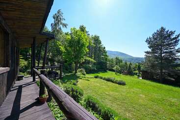 Un porche rústico de madera con vistas a un frondoso jardín, rodeado de árboles y colinas lejanas bajo un cielo azul claro en un día soleado.