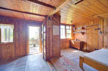 El interior de esta rústica cabaña de madera cuenta con grandes ventanas, una puerta abierta con vistas al exterior, una cama sencilla, acogedoras alfombras, luz natural y una encantadora decoración vintage en todas partes.