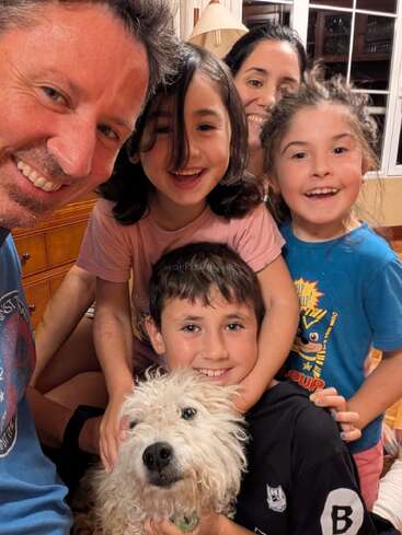Un selfie de famille plein de joie, avec cinq personnes souriantes et un chien blanc tout doux. Tout le monde a l'air heureux et complice, immortalisant un moment chaleureux, amusant et plein d'affection, pris à l'intérieur.