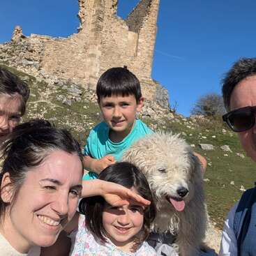 Une famille avec deux enfants et un chien blanc tout doux sourit pour un selfie pris en extérieur par une journée ensoleillée, avec des ruines de pierre anciennes en arrière-plan.