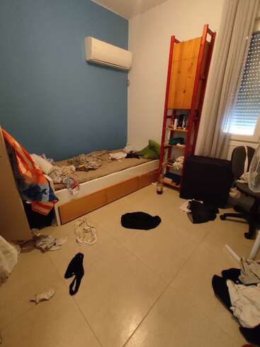 Esta imagen muestra una habitación desordenada con ropa esparcida por el suelo, una cama sin hacer, una estantería roja con escalera, una ventana y un aire acondicionado.