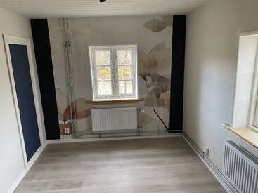 This image shows a small, empty room with light wood flooring, two windows, radiator, floral wall mural, white walls, and black vertical accents near the corners.