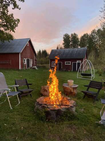 A cozy backyard scene with a blazing campfire surrounded by chairs, rustic wooden buildings, green grass, swing, trees, and a soft colorful evening sky.