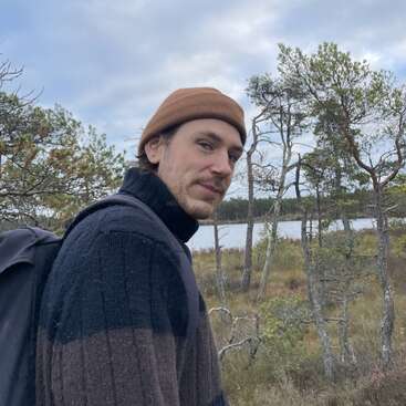 A man wearing a brown beanie, dark sweater, and backpack stands outdoors near leafless trees and a lake under a cloudy sky, enjoying nature.