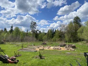 A grassy yard with garden beds, a lawn mower, and rocky piles. Surrounded by trees under a blue sky filled with fluffy white clouds. Peaceful atmosphere.