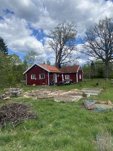 A quaint red house with a tiled roof sits among green grass, garden beds, and trees under a partly cloudy sky in a peaceful countryside setting.