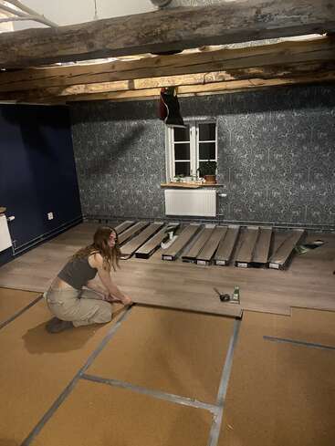 Uma mulher está colocando piso laminado em um cômodo aconchegante com vigas de madeira à vista, paredes com papel de parede e uma janela com vasos de plantas no parapeito.