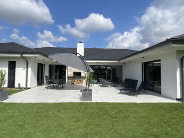 Ein modernes weißes Haus mit einer geräumigen Terrasse mit Liegestühlen, Essplätzen im Freien, einer Feuerstelle, einem großen Sonnenschirm, grünem Gras, blauem Himmel und vereinzelten Wolken darüber.