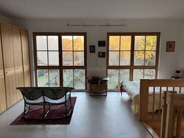 This cozy room features large windows overlooking a scenic outdoors, a wicker bench on a red rug, a wooden wardrobe, a single bed, and minimal decor.