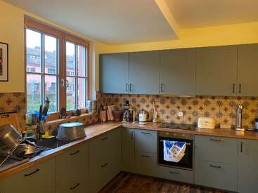 A cozy, L-shaped kitchen with green cabinets, wood countertops, floral tile backsplash, yellow walls, large window, and various appliances. Dishes are drying beside the sink.