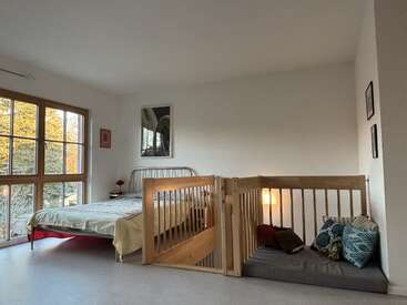 A cozy bedroom with large windows, a metal bed, light bedding, bedside table, framed art, and a wooden enclosed play area with cushions and a small lamp.