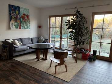 This cozy living room features a gray sofa, wooden coffee table, modern chair, large windows, indoor plant, colorful artwork, natural light, and a warm atmosphere.