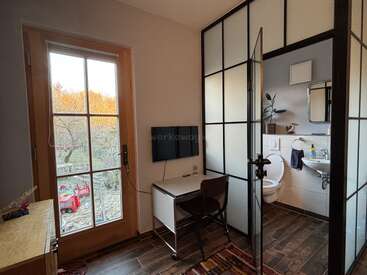 This image shows a small, modern room with a desk, chair, TV, and a glass door. The adjoining bathroom is visible, featuring a toilet and sink.