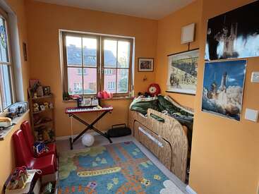 This is a cozy children's bedroom with orange walls, a wooden bed, colorful carpet, musical keyboard, toys, posters, and large windows letting in daylight.