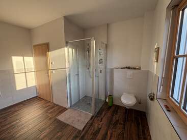 This image shows a modern bathroom with a glass shower, floating toilet, wooden floor tiles, large window, beige walls, and a door. Light fills the space.