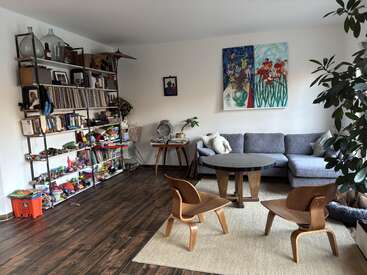 A cozy living room with wooden floors, a grey sectional sofa, colorful wall art, shelves filled with books and toys, two wooden chairs, and plants.