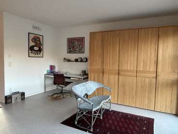 A cozy room with a wooden wardrobe, a simple desk with chair, wicker bench on a red rug, and colorful art on the white walls.