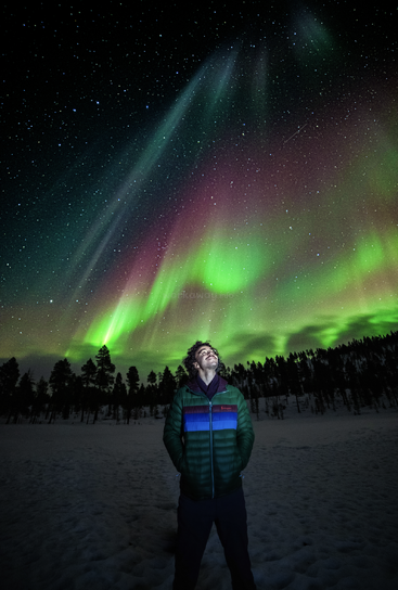 The image depicts a man standing in a snowy landscape, gazing upwards at the night sky, where the aurora borealis is visible in shades of green and red.